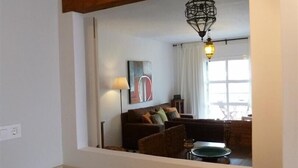 A kitchen with a wooden beam ceiling, a dining table with chairs, a hanging light fixture, and a view into a living room with a sofa and a painting. - One of the best located apartments in Conil.Sea views from every room. With WiFi (Conil de la Frontera)