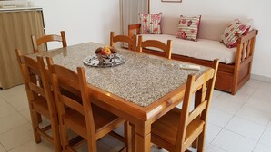A dining area with a granite table, wooden chairs, and a fruit bowl. There is a sofa with patterned cushions in the background.