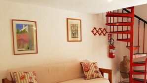 A cozy living room with a wooden sofa, two cushions, a framed picture, a small table with a vase, and a red spiral staircase.