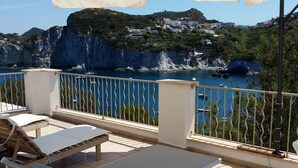 A balcony with a white umbrella, two lounge chairs, and a view of a coastal area with cliffs and buildings.