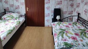 Una stanza modesta con due letti singoli, una porta in legno e un tavolino con una lampada.