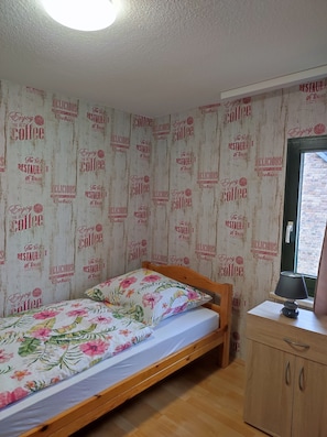 A small bedroom with a single bed, wooden headboard, a nightstand with a lamp, and a wardrobe. The walls have a vintage coffee-themed wallpaper.