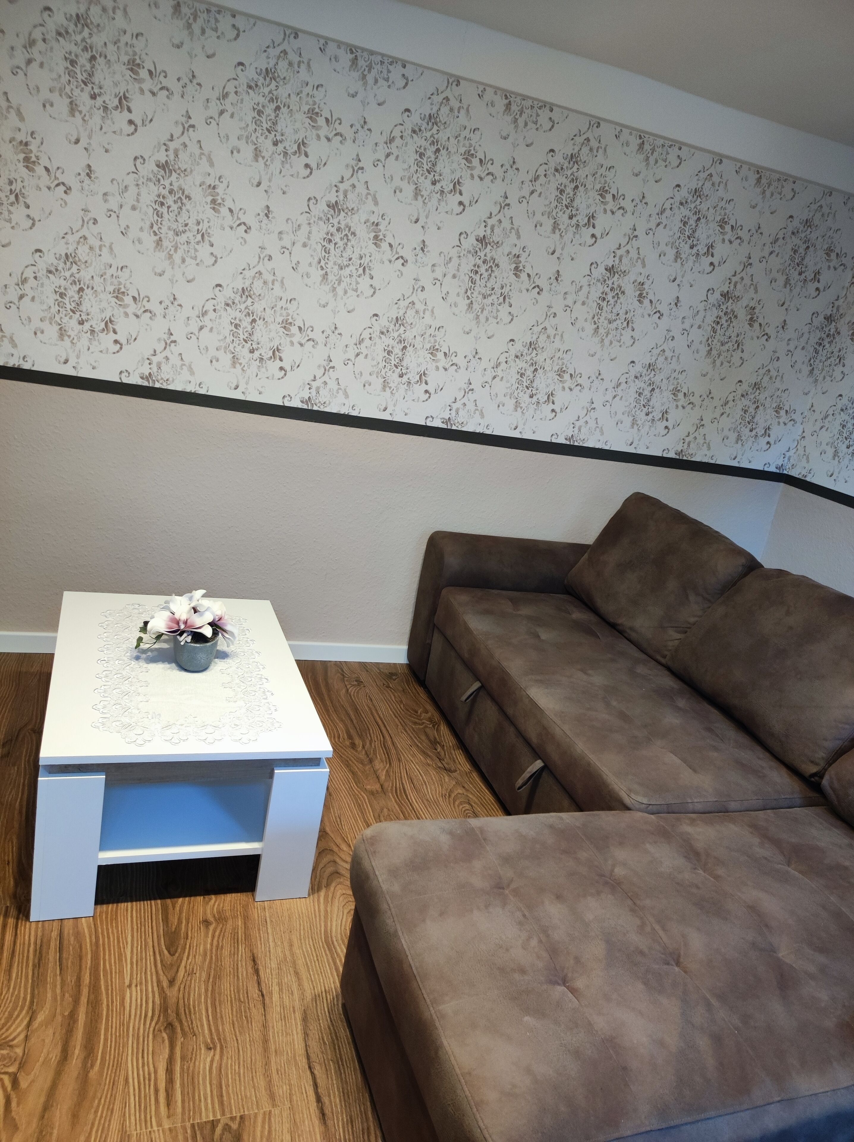 Una habitación con una mesita de noche blanca, un sofá marrón y un papel tapiz floral.