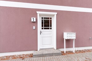 Eine weiße Tür mit Fensterglas, ein Briefkasten und eine Beschilderung mit der Nummer 1 an einer rosafarbenen Wand.