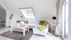 Uma sala de estar moderna com um sofá branco, uma mesa de centro e uma pequena mesa lateral. Há um claraboia acima do sofá, uma janela à direita e uma porta à esquerda.