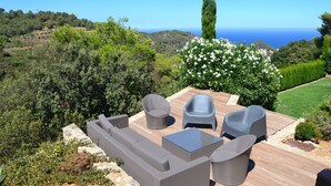 Una terraza con mobiliario exterior, vista al mar y un jardín con un árbol alto y flores blancas.