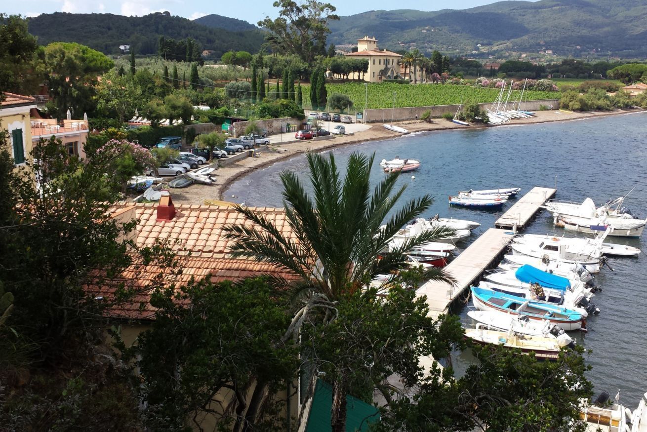 A marina with boats docked, a building with a red-tiled roof, and a mountainous backdrop.