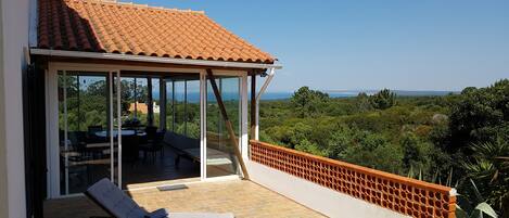 Terraço com duas espreguiçadeiras, porta de vidro que dá para uma área de refeições ao ar livre e vista para uma paisagem verdejante.