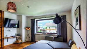 A bedroom with a bed, a TV, a lamp, and a window with a view of houses and mountains.