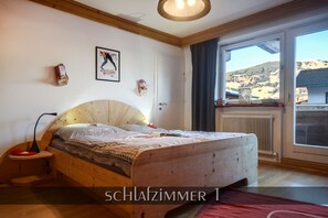Une chambre à coucher avec un lit en bois, une table de chevet, une lampe, un tableau encadré au mur et une fenêtre donnant sur la campagne.