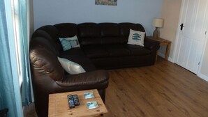 A living room with a brown leather sofa, a small wooden coffee table with remotes, a lamp, and a painting on the wall. - Dog Friendly, Cosy Fisherman's Cottage Within 150 Metres Of A Long Sandy Beach  (Caister-on-Sea)