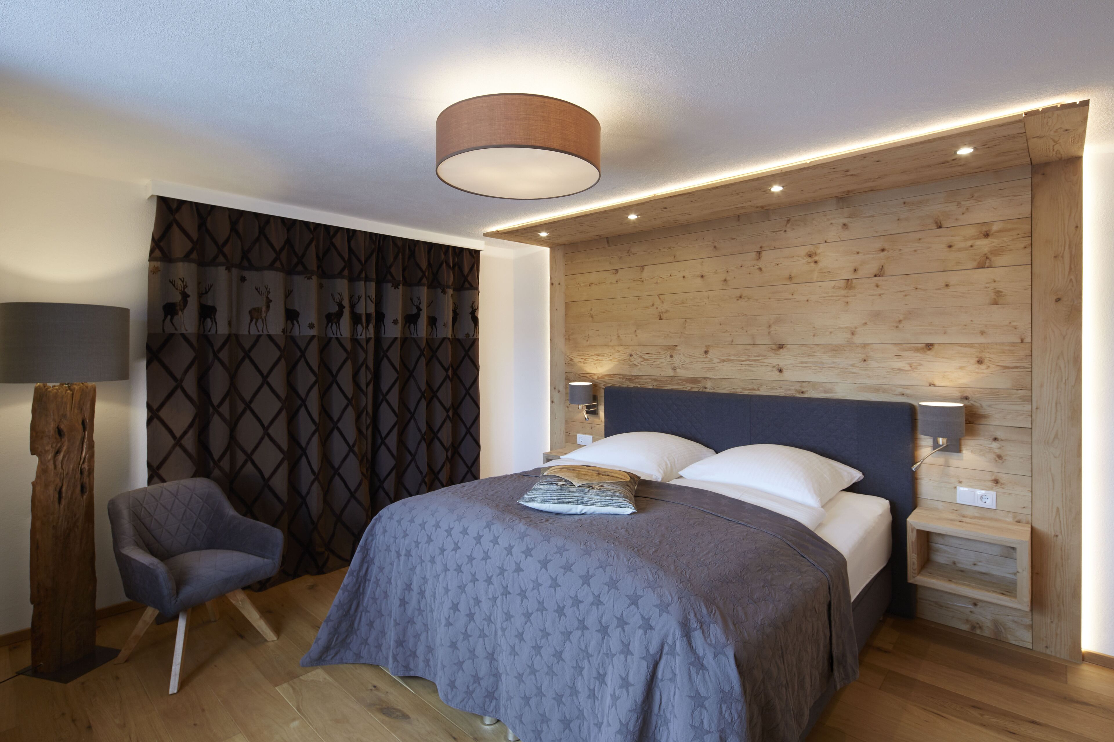 A modern bedroom with a wooden headboard, a bed with a patterned bedspread, a nightstand with a lamp, and a chair.