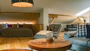 Un accogliente soppalco con camera da letto, soffitto a falde, travi in legno, un letto con biancheria a motivi decorativi, un divano grigio e un tavolino in legno.