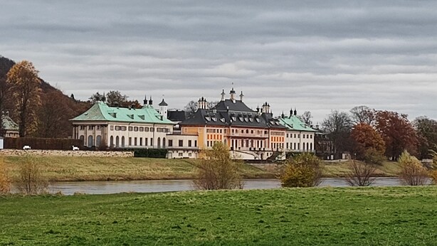 A large, historic building with multiple wings and green roofs, surrounded by a grassy field and a river.