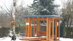 A wooden gazebo with a bench, surrounded by snow-covered ground and trees.