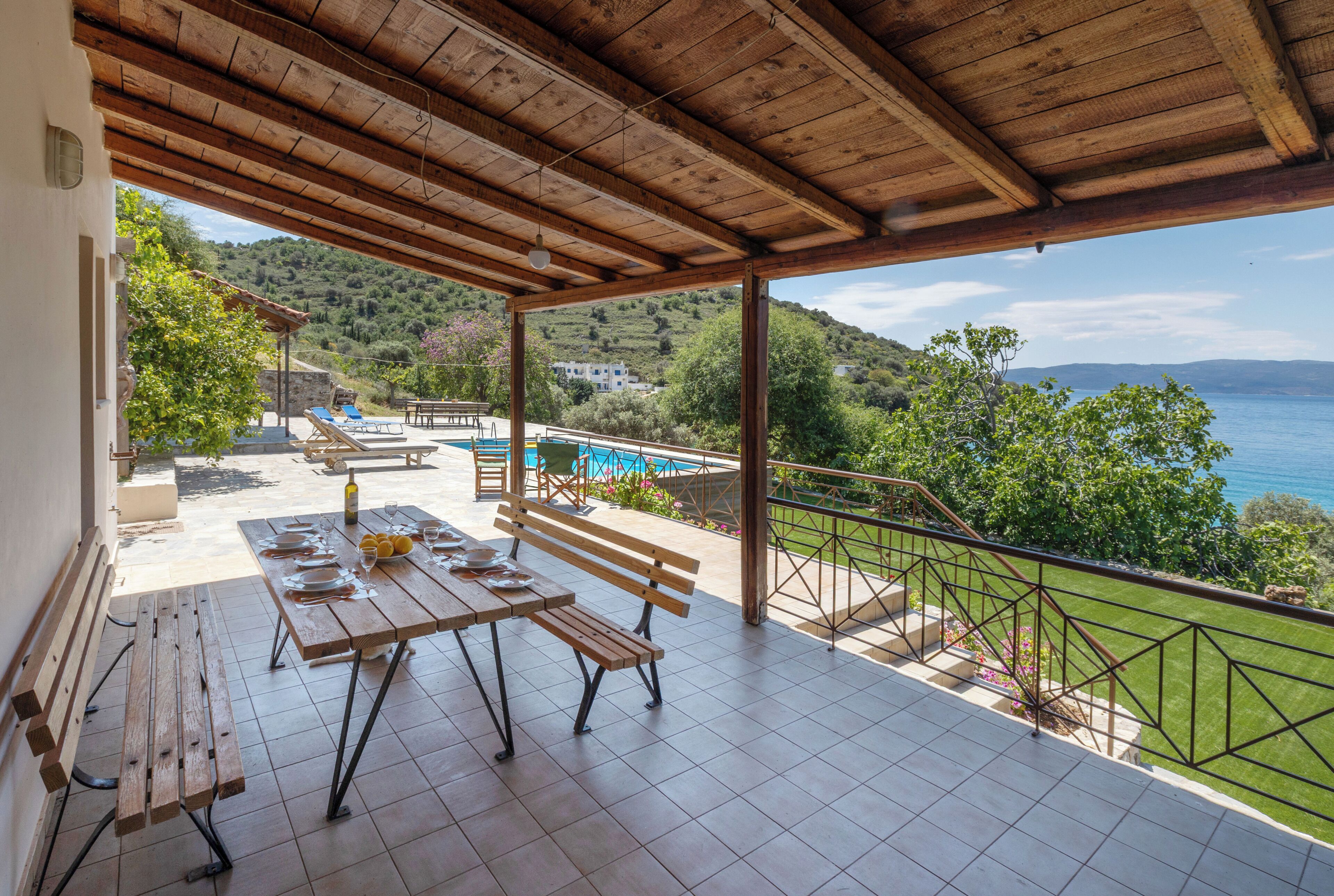 A covered outdoor patio with a pool, wooden furniture, and a scenic view of the sea.