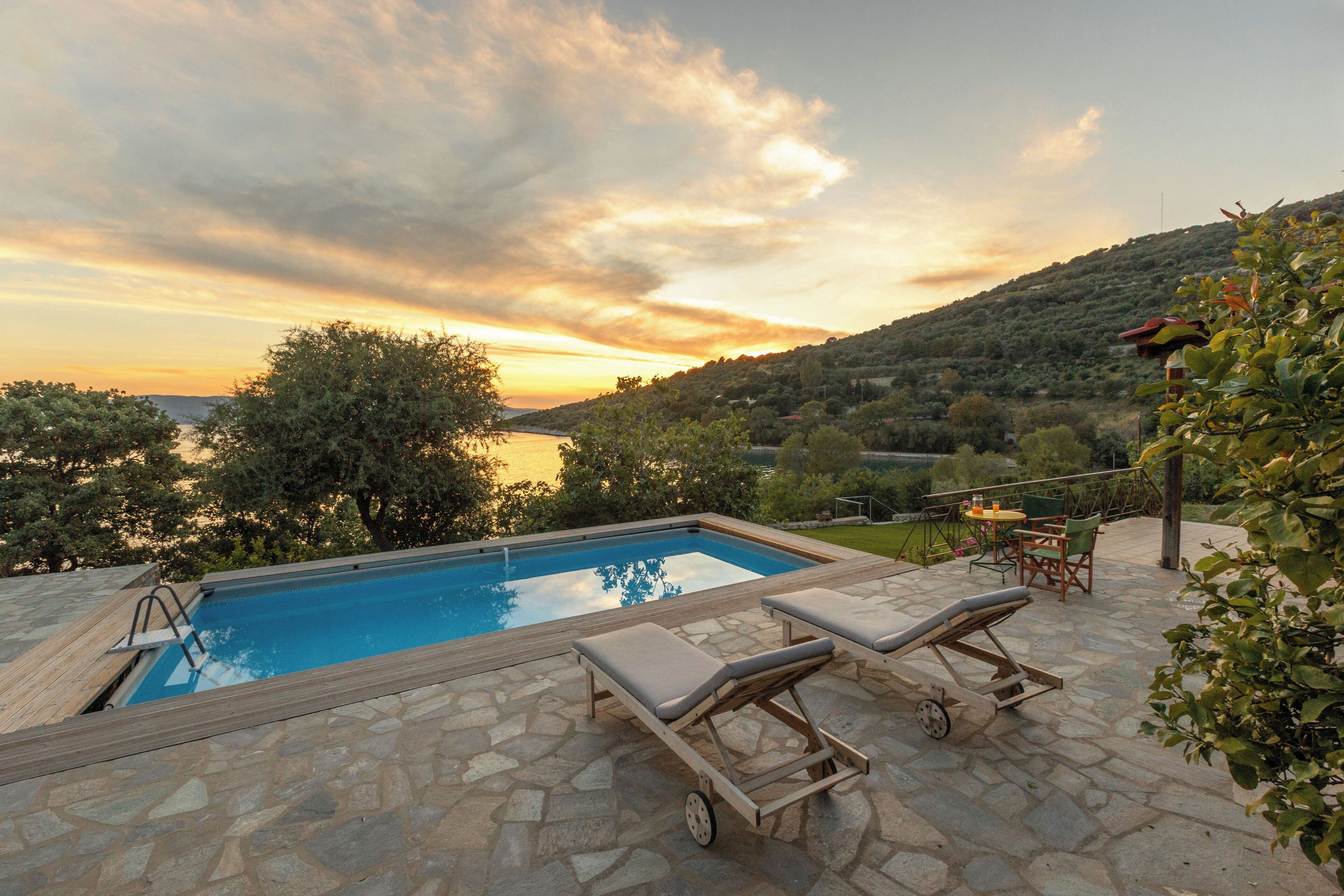 A pool with lounge chairs and a table, surrounded by stone tiles and greenery.