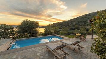 A pool with lounge chairs and a table, surrounded by stone tiles and greenery.