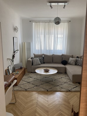 Un salon moderne avec un parquet en bois disposé en chevrons, un canapé d’angle beige, une table basse blanche et une grande fenêtre agrémentée de rideaux légers.