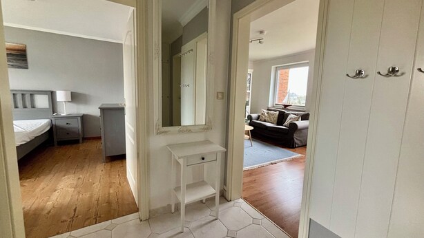 Een gang die leidt naar een slaapkamer met een bed, nachtkastje en lamp. Er hangt een spiegel aan de muur, en er is een deur naar een andere kamer.