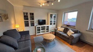 Een woonkamer met een grijze bank, een kleinere grijze bank, een houten salontafel en een televisie op een wit kastje. Er is een raam met uitzicht op een gebouw en een lamp op een bijzettafeltje.
