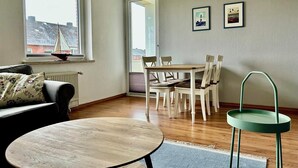 Een gezellige woonkamer met een ronde houten tafel, een zwarte bank en een eethoek met een witte tafel en stoelen.