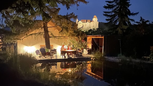 A lakeside outdoor seating area with a table and chairs, surrounded by trees and a castle in the background. - Organic home, garden & swimming pond, quiet location, Vienna reached in 15 min (Maria Enzersdorf)
