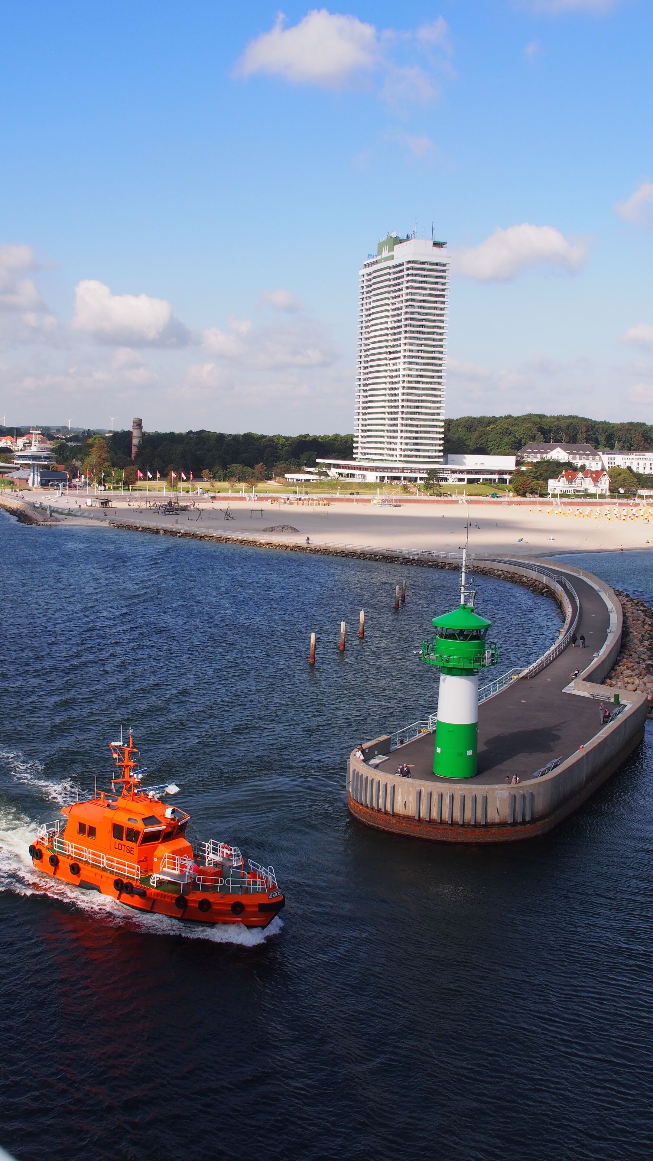 Eine Küstenansicht mit einem großen, orangefarbenen Schlepper, einem grün-weißen Leuchtturm und einem hohen, weißen Gebäude im Hintergrund.