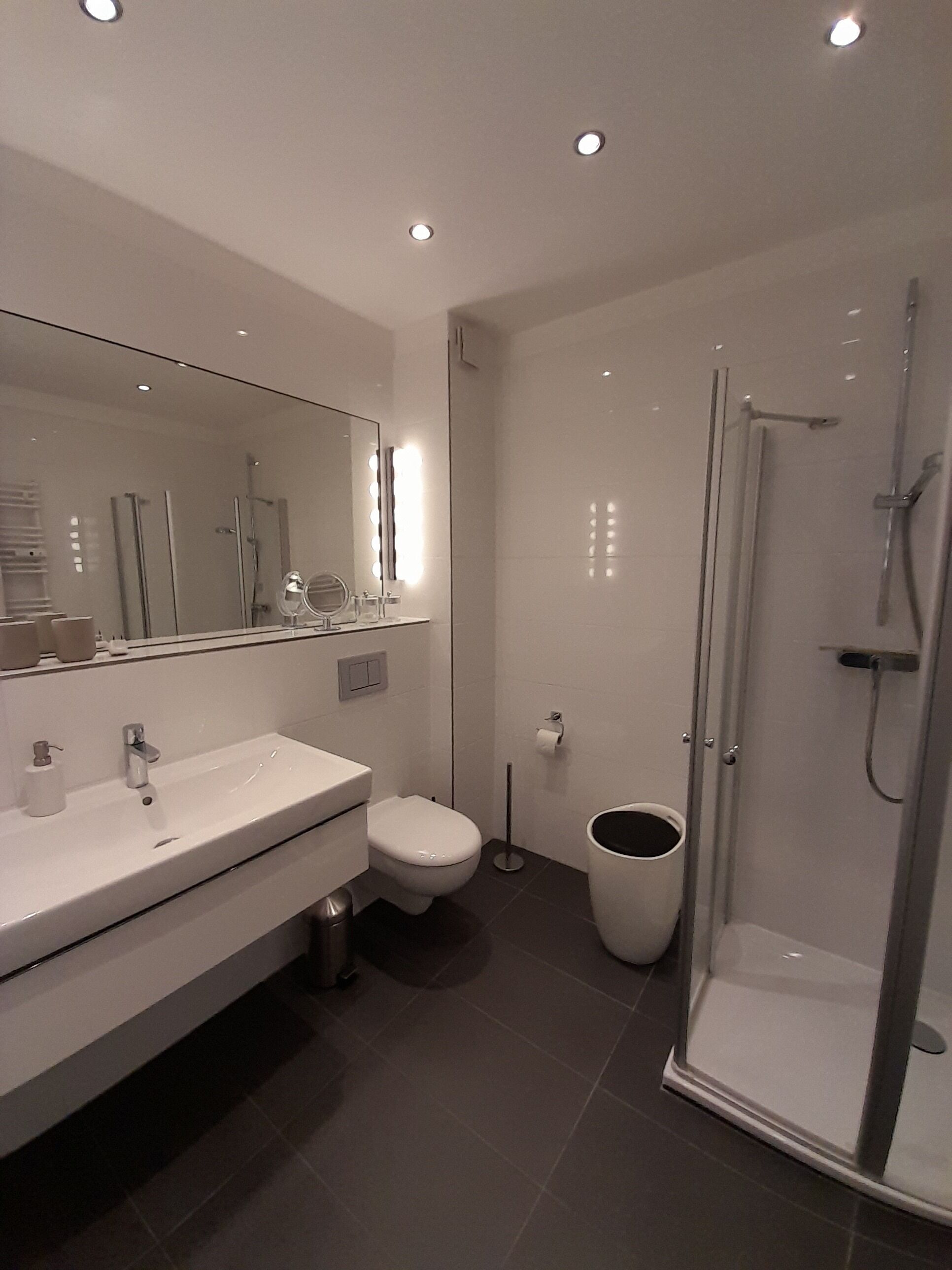 Une salle de bain moderne avec un meuble-lavabo blanc, des toilettes, une douche à parois de verre et un grand miroir avec éclairage intégré.