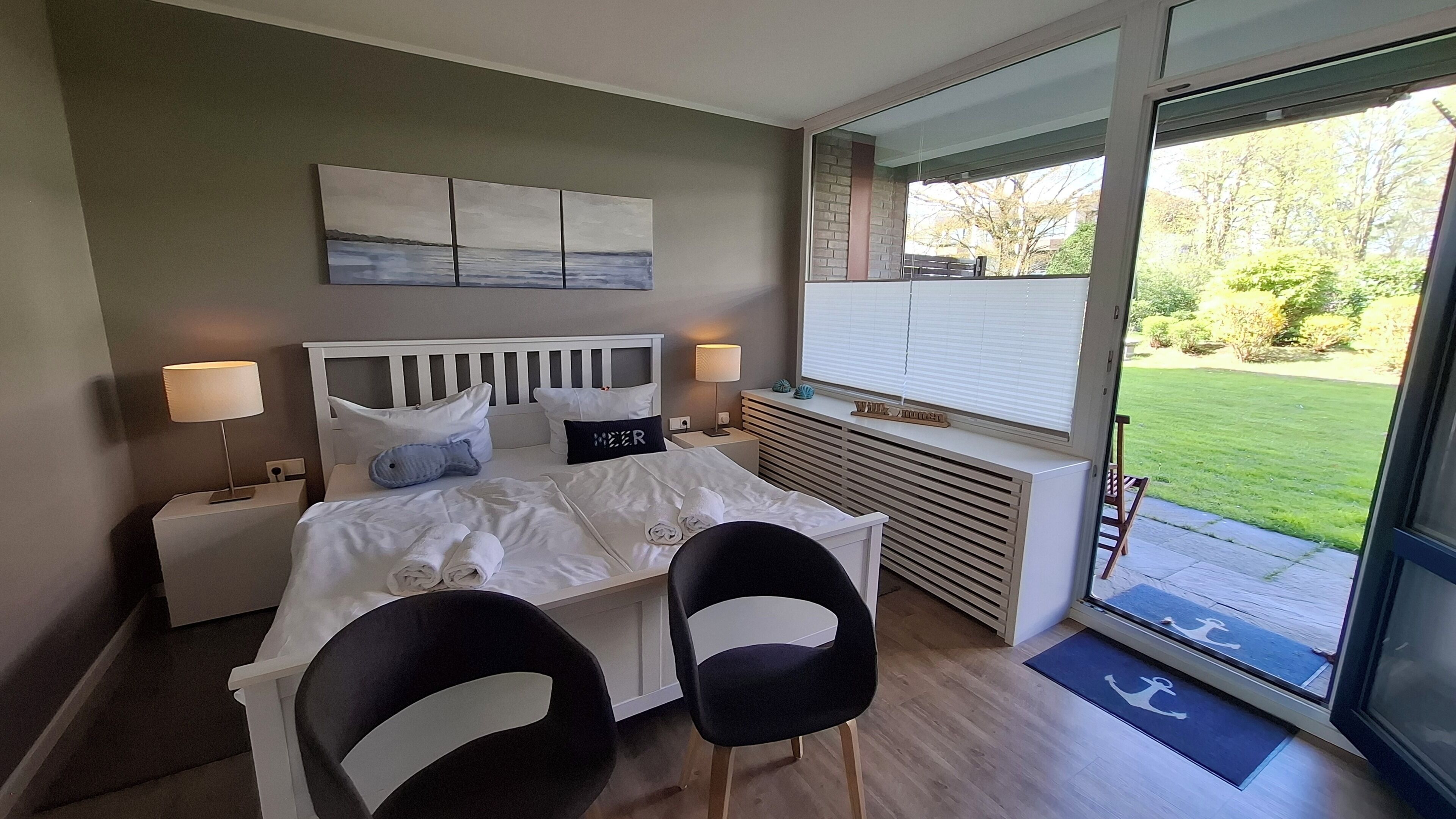 A modern hotel room with a bed, two chairs, a nightstand, a lamp, a window with a view of the outdoors, and a wall-mounted artwork.