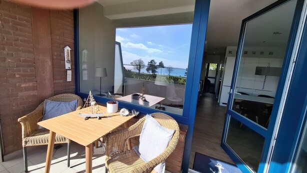 A balcony with a wooden table and wicker chairs, a view of the sea, and a glass door leading to a kitchen.