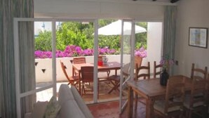 Quarto com teto de madeira, porta de vidro deslizante que dá para área externa com mesa e cadeiras, e vista para vegetação e flores.