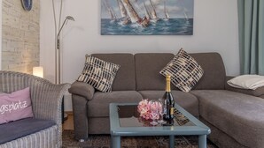 Een woonkamer met een bank, een salontafel met een fles en glazen, een vloerlamp en een schilderij van zeilboten.