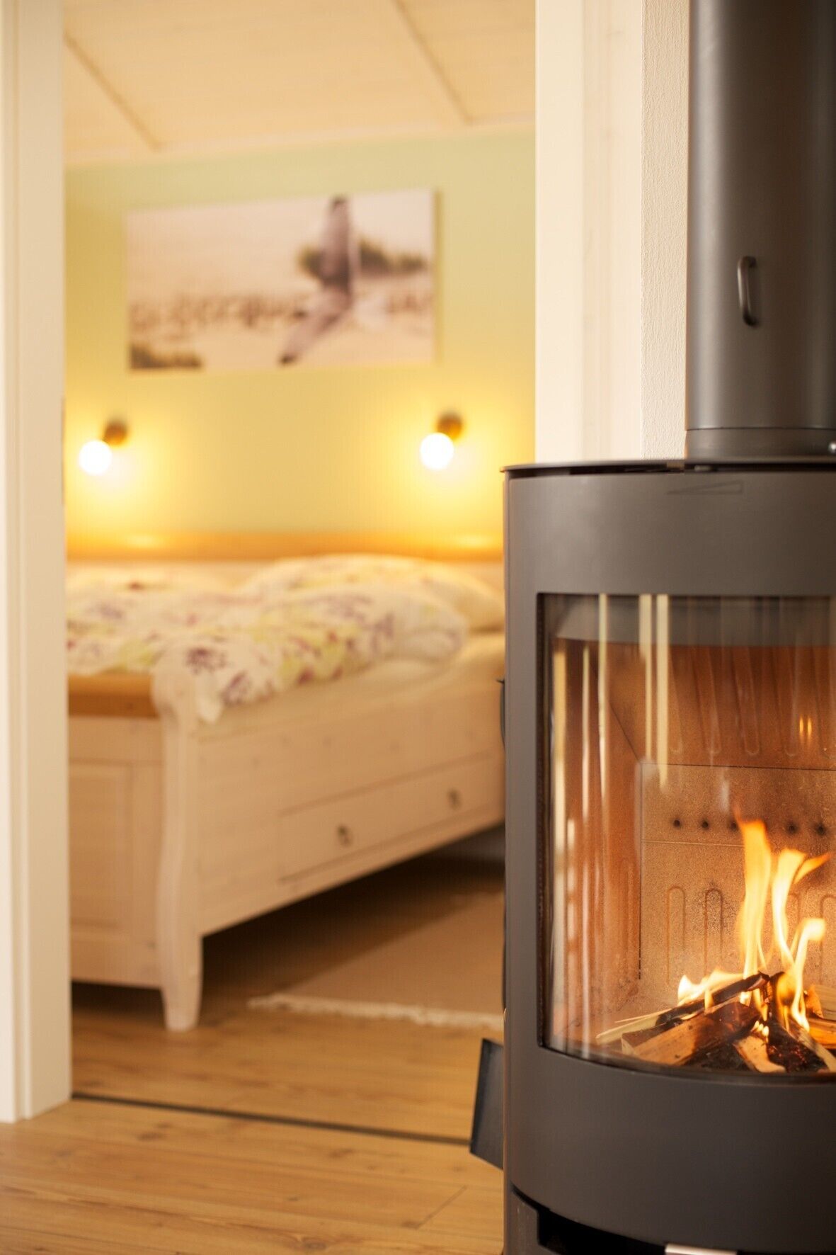 A room with a bed, a wall-mounted heater, and a visible fire burning inside a stove.