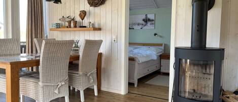A cozy room with a dining area, wicker chairs, a wooden table, a bed with a white headboard, a wall-mounted shelf with decorative items, a fireplace, and a door leading to another room.