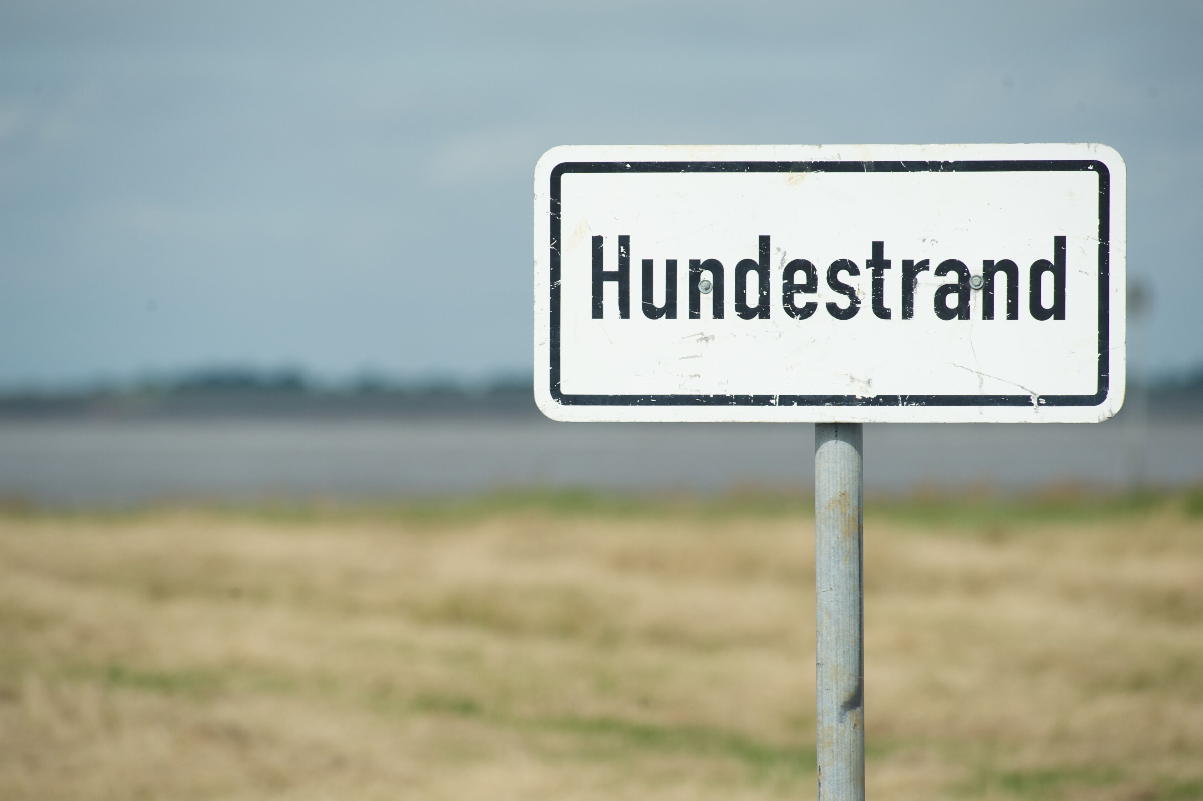 Un letrero con el texto 'Hundestrand' sobre un fondo borroso de un campo y una masa de agua.