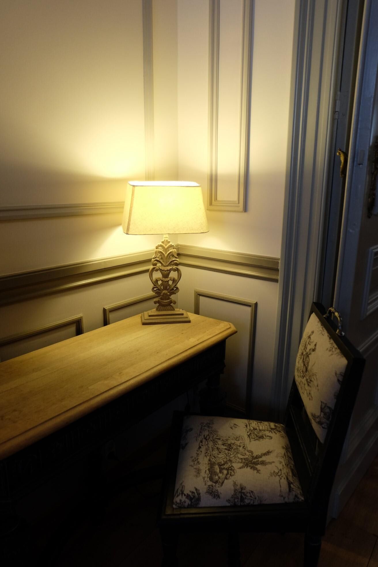 A corner room with a wooden table and a chair, a lamp on the table, and a door with a handle.
