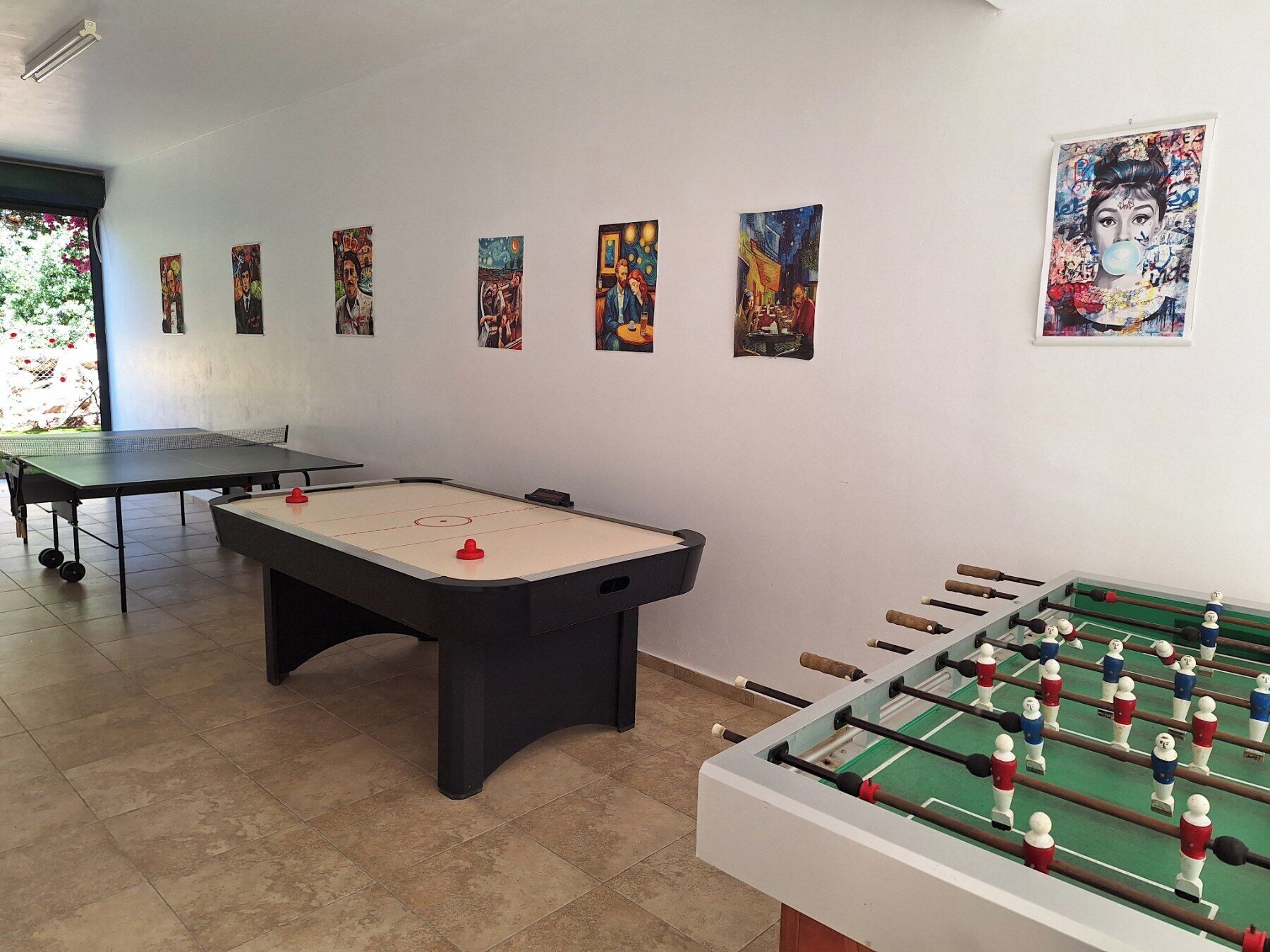 A room with a foosball table, a ping pong table, and several artworks on the walls.
