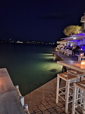 Eine Bar am Wasser mit Außenbereich, Blick auf das Wasser und einer Stadtkulisse im Hintergrund.