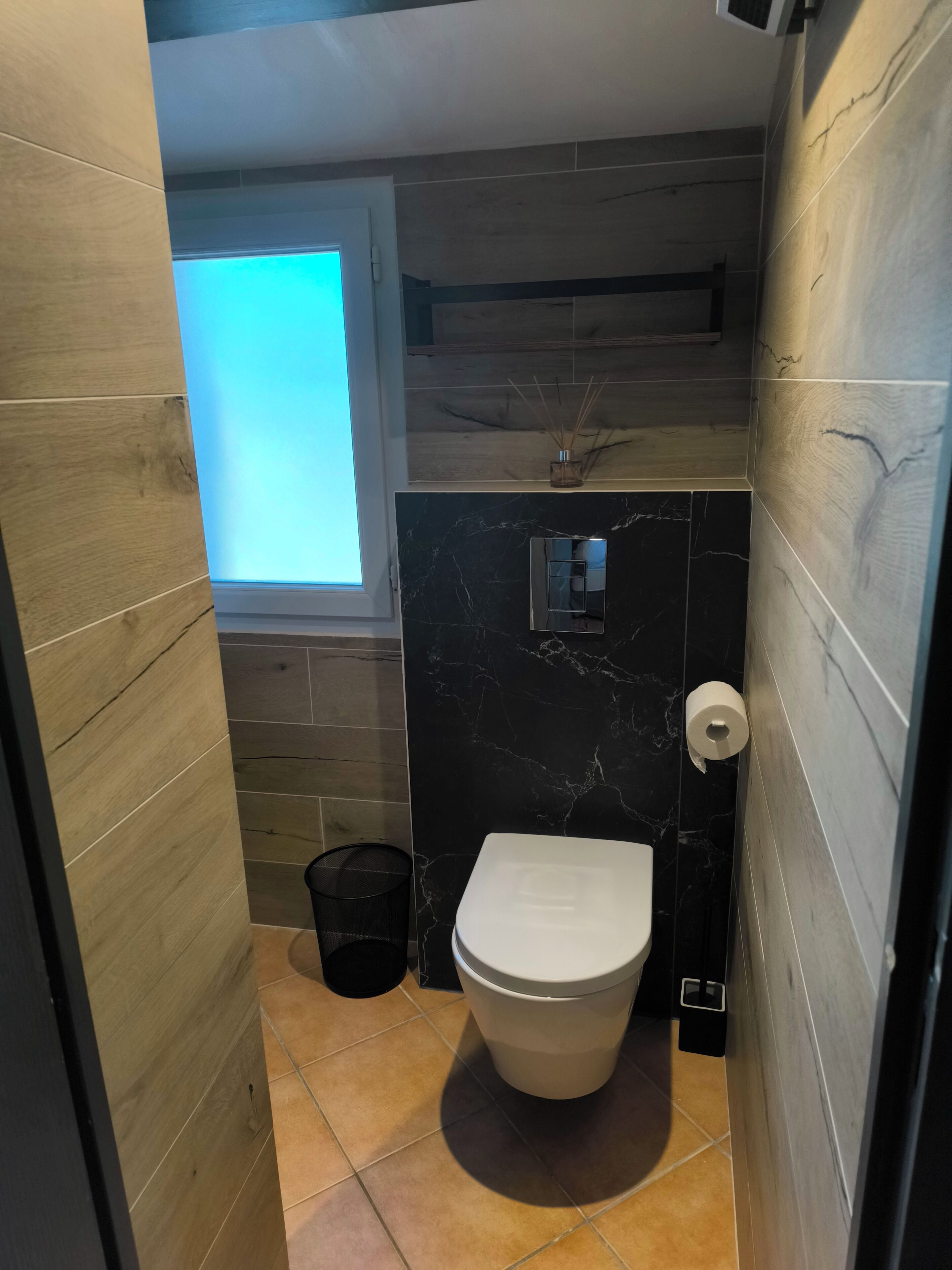 A compact bathroom with a toilet, a trash bin, a window, and a shower area.