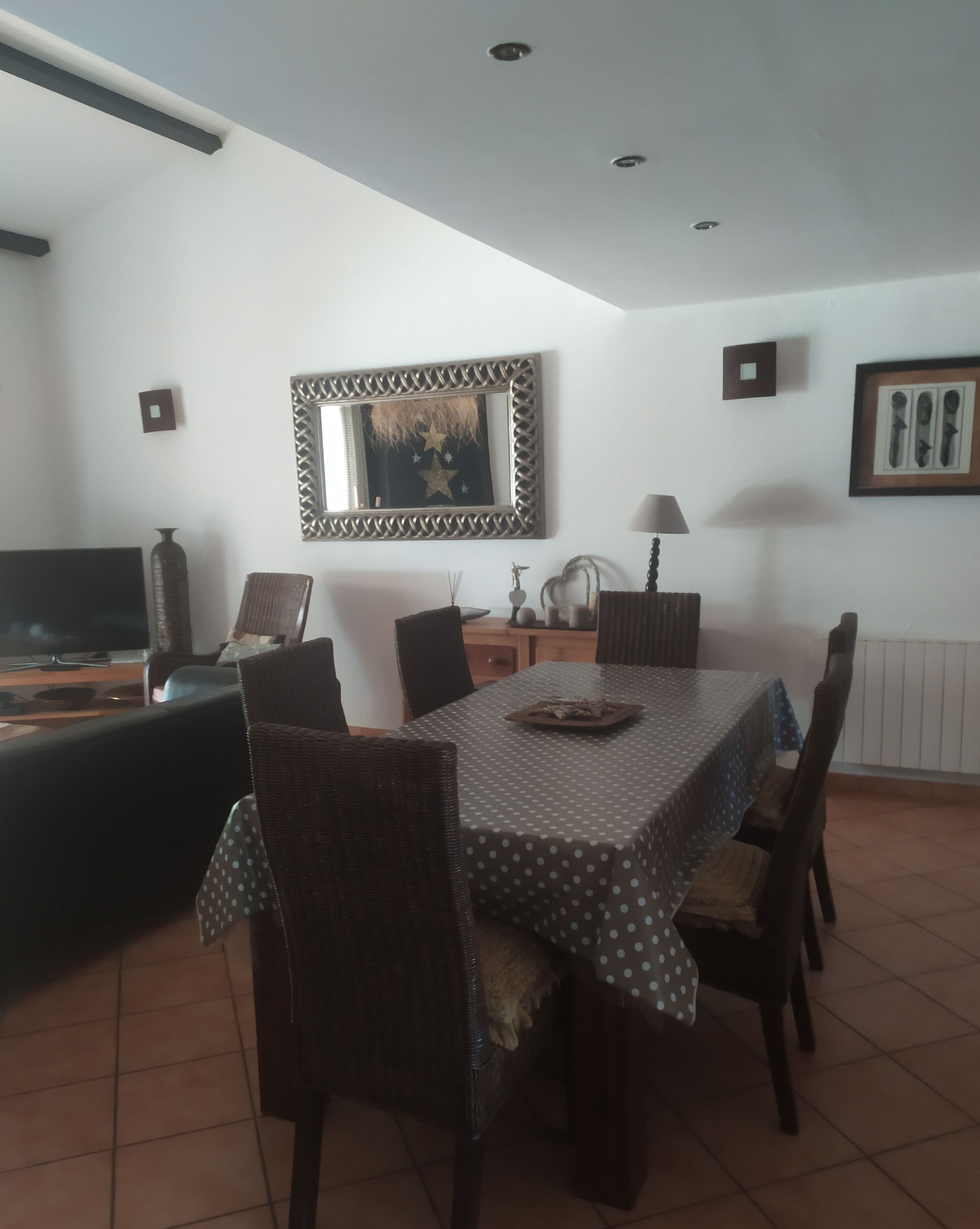 A dining area with a table covered in a polka dot tablecloth, surrounded by wicker chairs. A television is visible in the background.