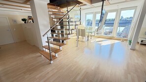Un intérieur moderne avec un escalier en bois, un coin repas et de grandes fenêtres offrant une vue sur l’extérieur.