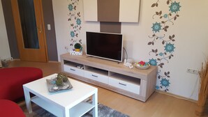 A living room with a TV on a wooden stand, a small table with decorative items, and a red sofa. - Family-friendly apartment in the heart of East Frisia incl. Wireless Internet access (Westerholt)
