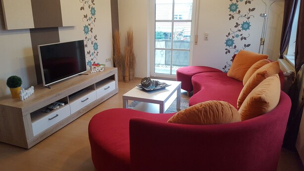 A living room with a flat-screen TV on a wooden TV stand, a red sofa with cushions, a white coffee table, and a window with a view of the outside. - Family-friendly apartment in the heart of East Frisia incl. Wireless Internet access (Westerholt)