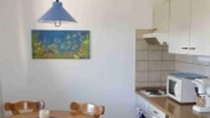 Een kleine, praktische keuken met witte kasten, een magnetron en een spoelbak. Er is een eettafel met stoelen en een blauwe hanglamp erboven.