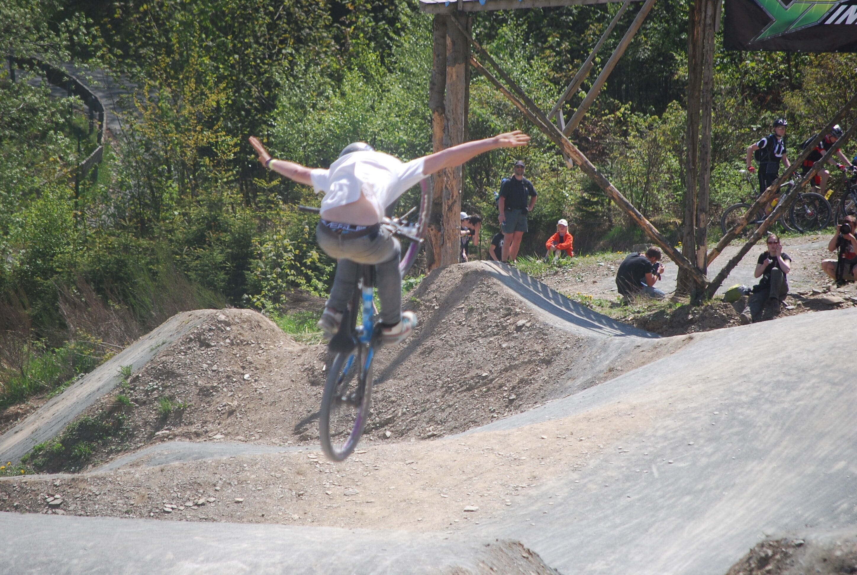 A person performing a BMX stunt on a dirt track with spectators and a photographer in the background.