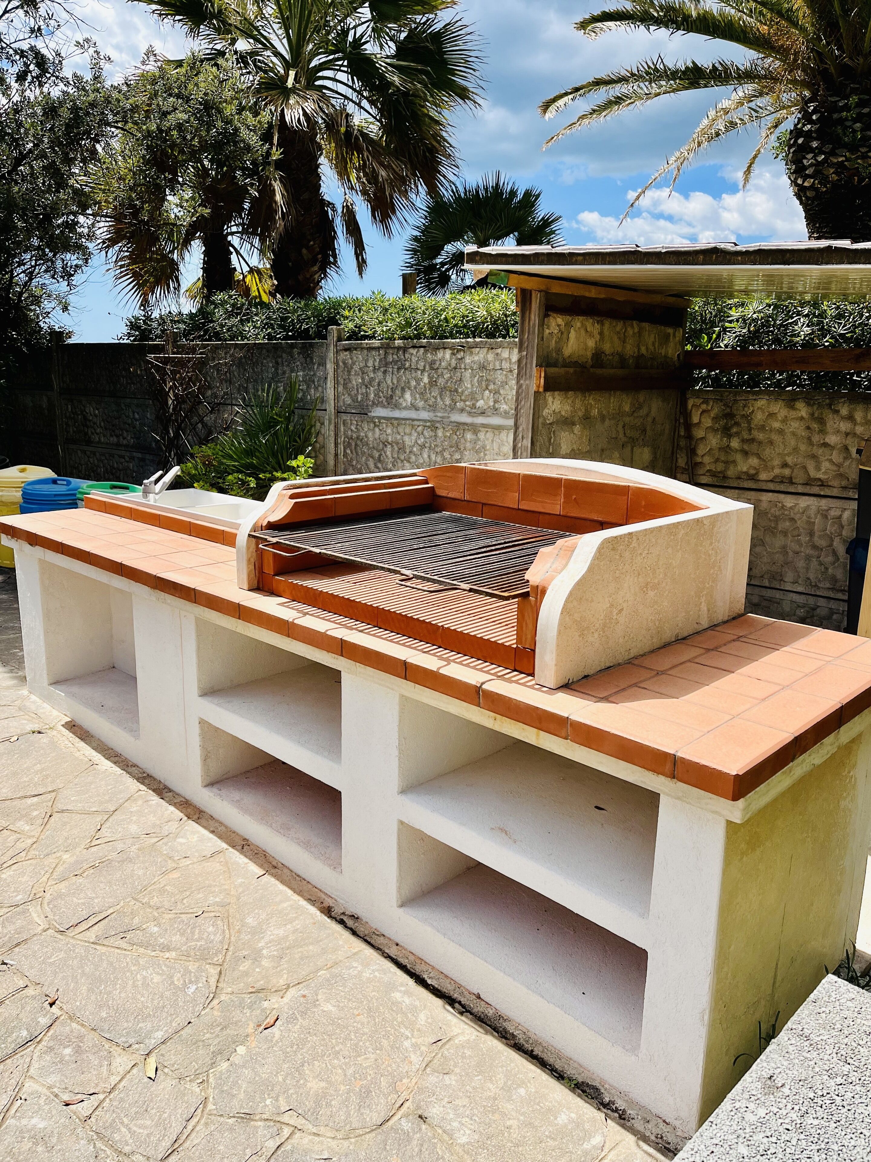 Un barbecue doté d’un évier intégré et d’étagères de rangement, installé devant un décor composé de palmiers et d’un mur en pierre.