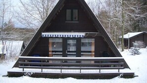 A triangular wooden cabin with a porch and windows, surrounded by snow and bare trees.