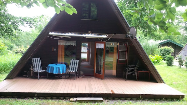 A triangular A-frame structure with a wooden deck, a table with chairs, and a glass door leading inside.