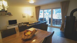 Een eetgedeelte met een houten tafel, stoelen en een schaal fruit. Er is een televisie, een bank en een open haard.